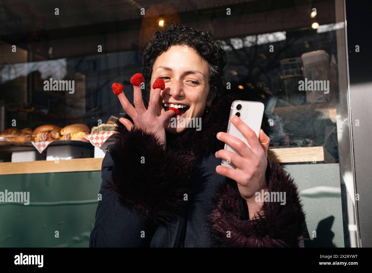 Donna giocosa che mangia lamponi con le dita e usa lo smartphone al caffè sul marciapiede Foto Stock