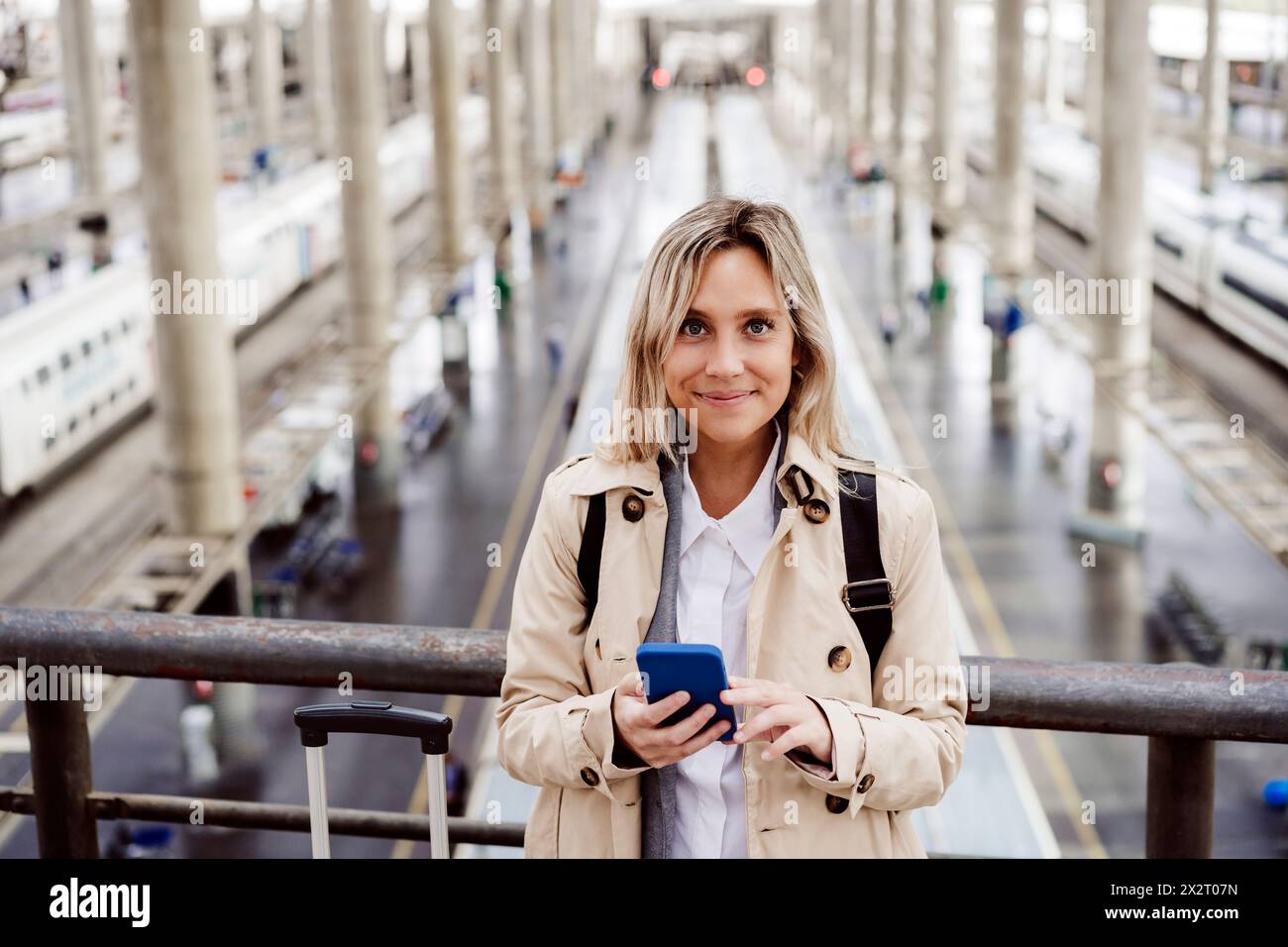 Donna sorridente con capelli evidenziati che tiene lo smartphone alla stazione ferroviaria Foto Stock
