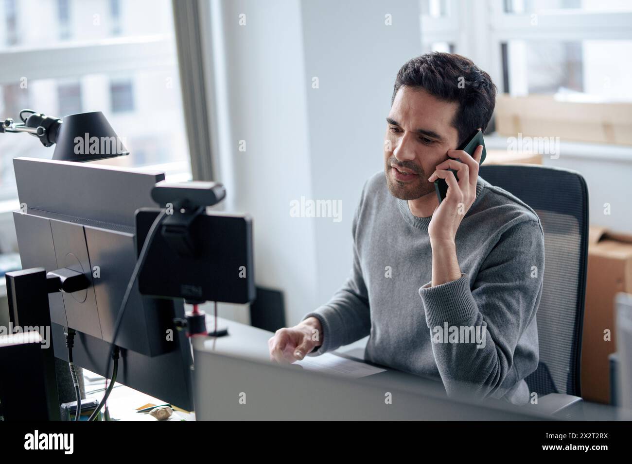 Uomo d'affari maturo che parla con uno smartphone vicino a un PC desktop alla scrivania Foto Stock