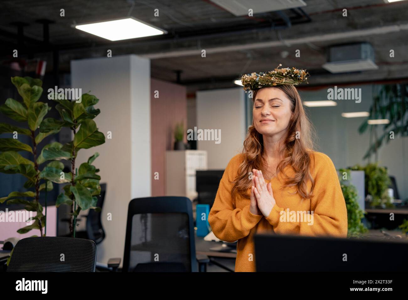 Donna d'affari sorridente con corona floreale e meditazione con le mani chiuse in ufficio Foto Stock
