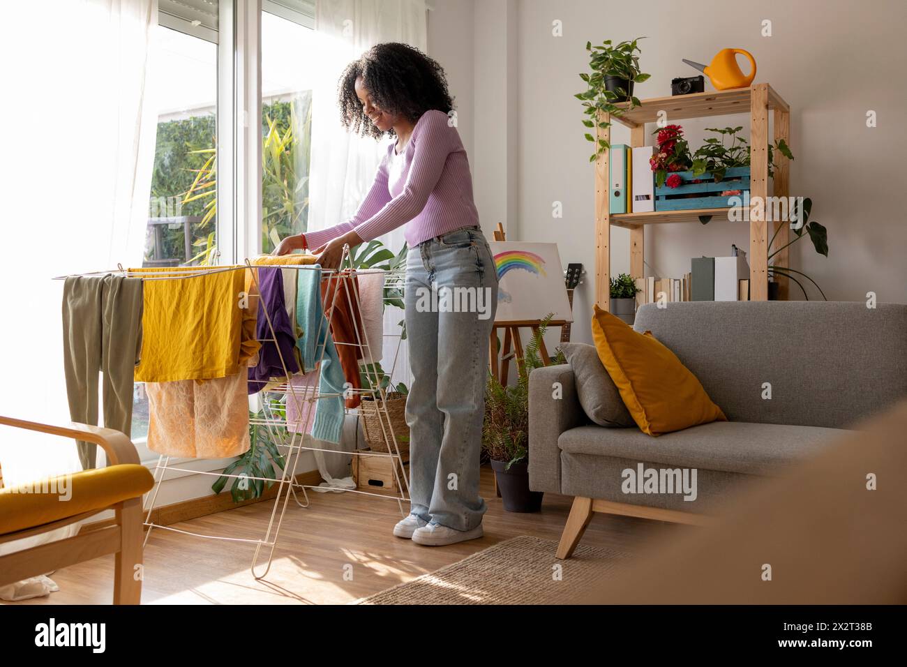 Giovane donna sorridente che asciuga i vestiti su un divano nel soggiorno Foto Stock