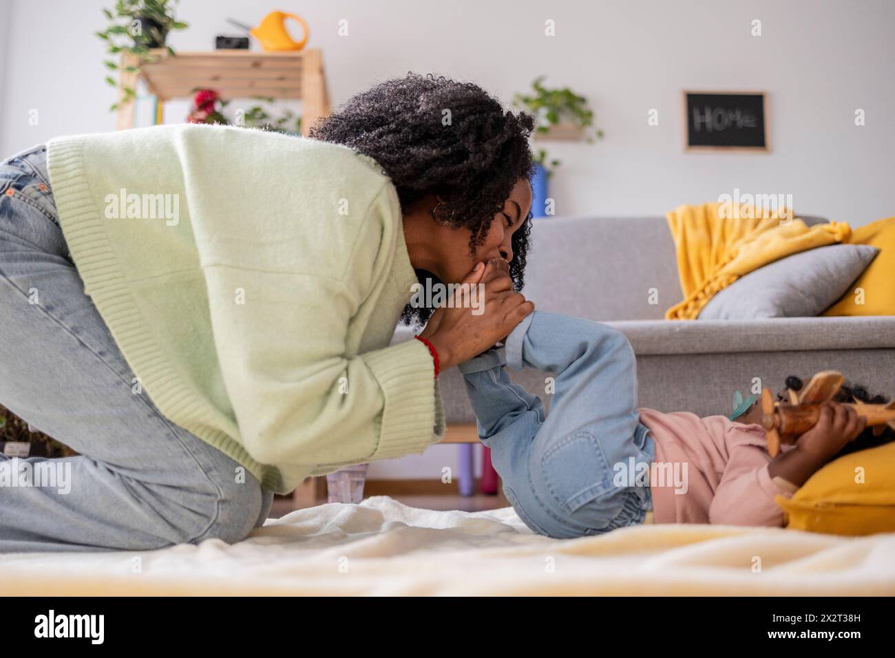 Giovane madre single che bacia il piede della figlia sdraiato con il giocattolo nel soggiorno di casa Foto Stock
