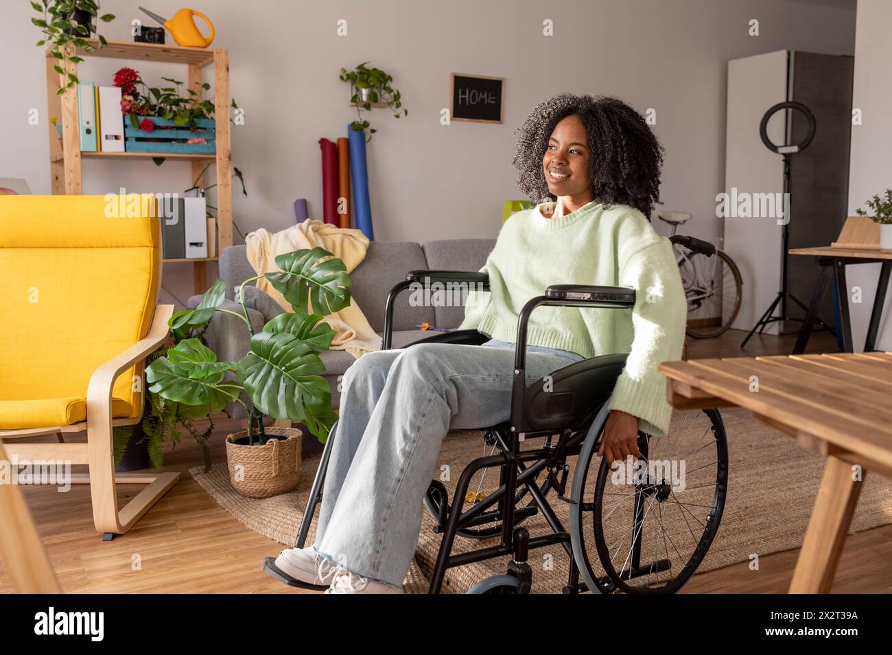 Giovane donna sorridente con disabilità seduta in sedia a rotelle a casa Foto Stock