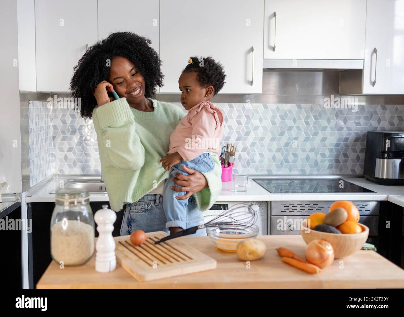 Madre single sorridente che porta la figlia che parla al telefono cellulare in cucina a casa Foto Stock