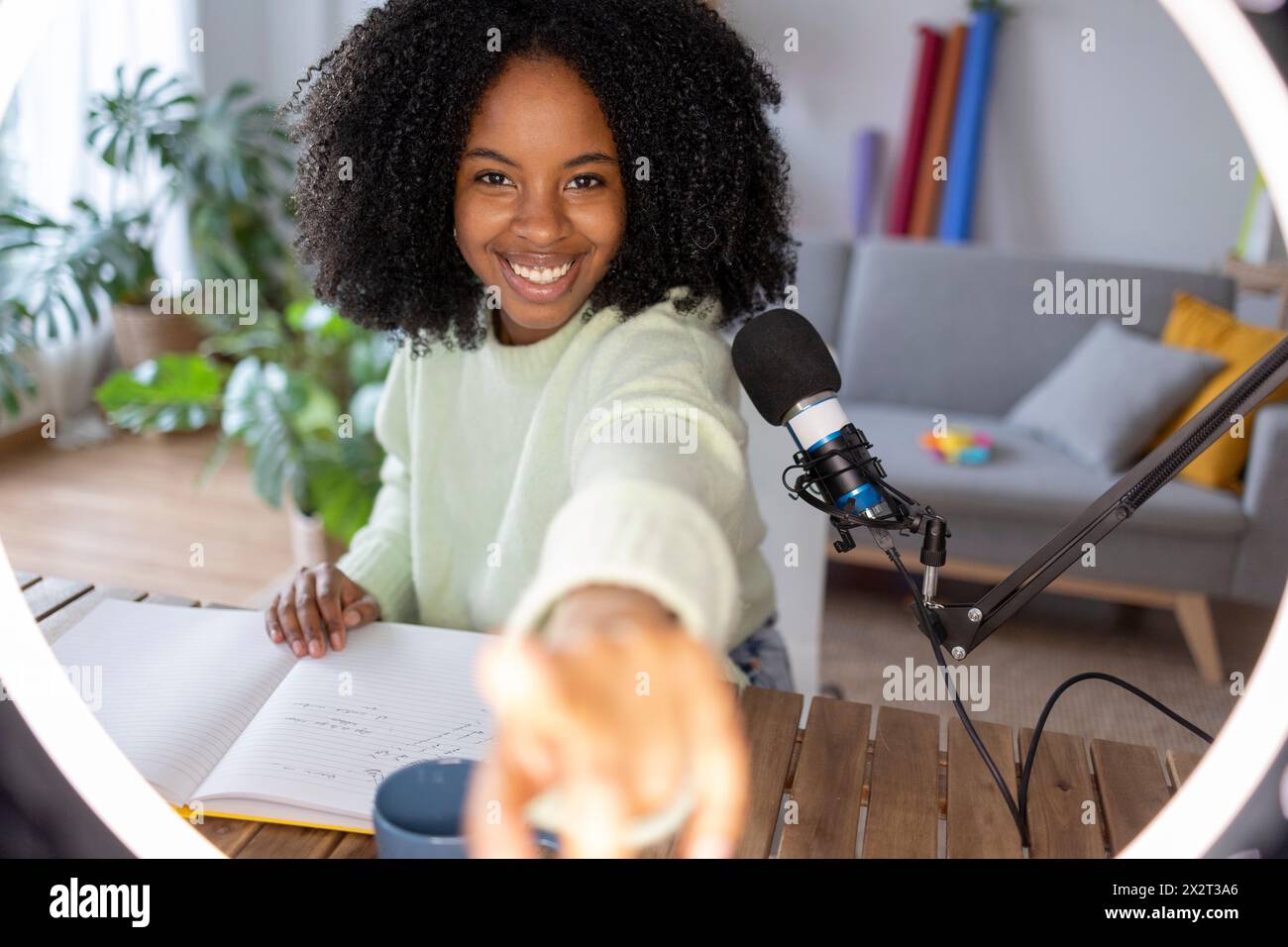 Giovane donna sorridente con microfono davanti alla luce anulare illuminata a casa Foto Stock