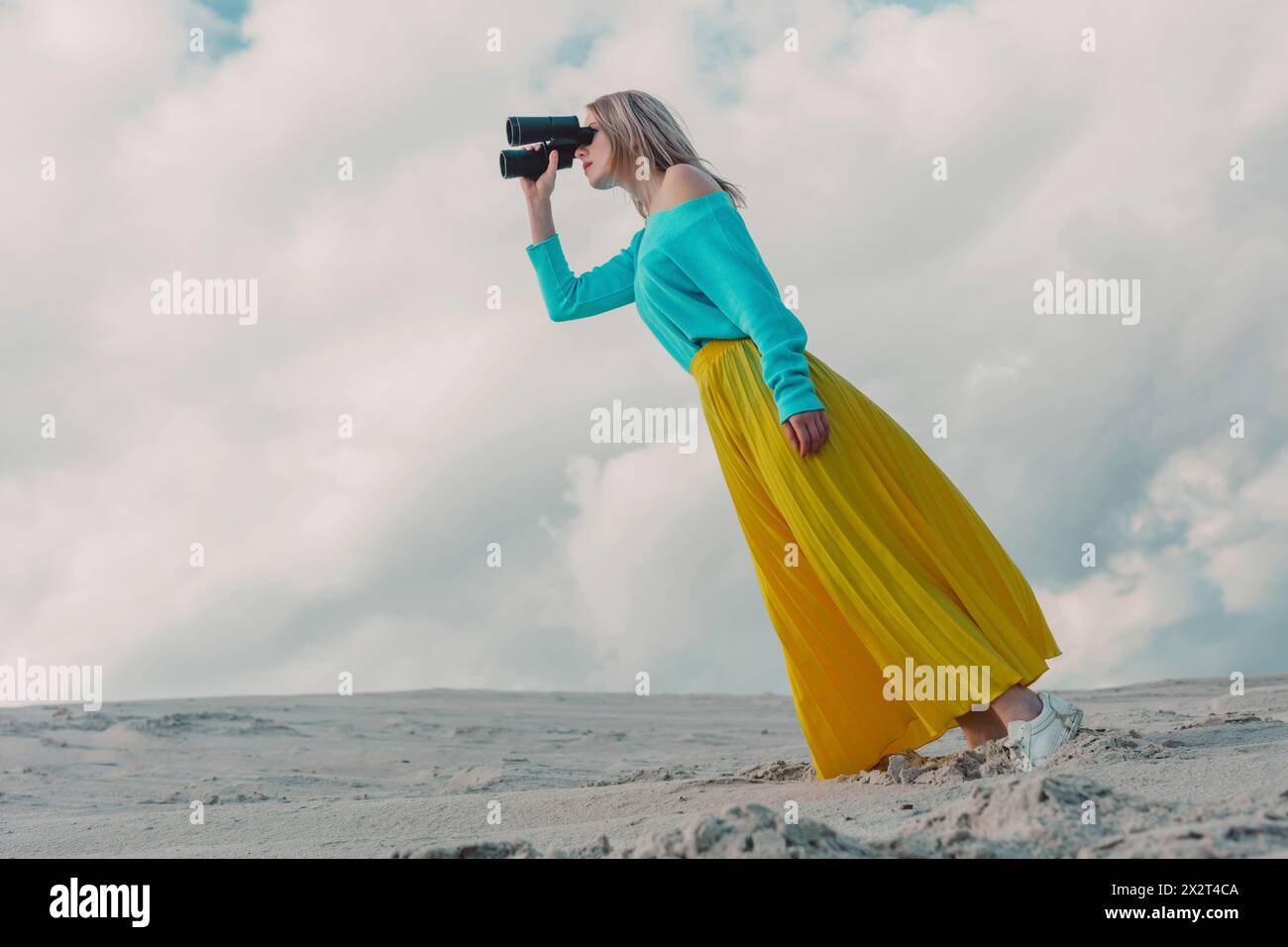 Donna che indossa una gonna gialla e guarda attraverso un binocolo sotto il cielo nuvoloso Foto Stock