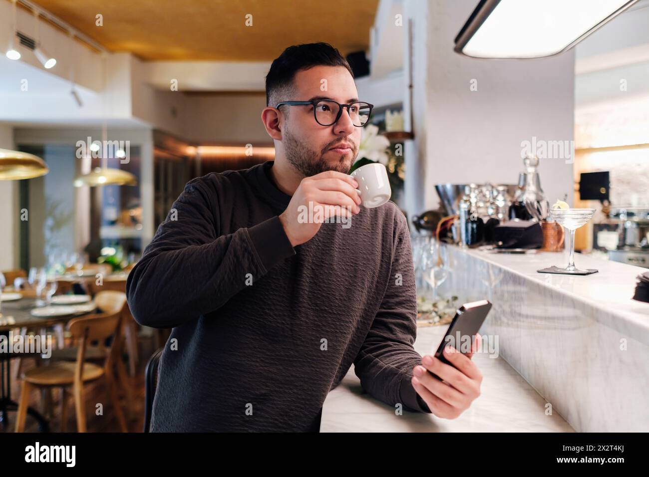 Cliente con telefono cellulare appoggiato al bancone del bar che beve caffè al ristorante Foto Stock