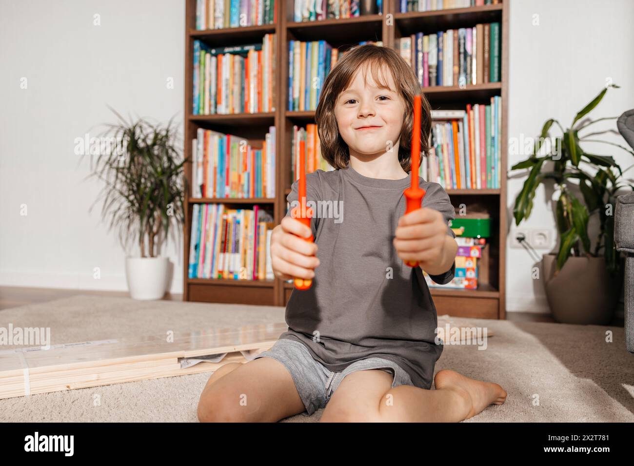 Sorridente che tiene i cacciaviti seduti davanti alla libreria a casa Foto Stock