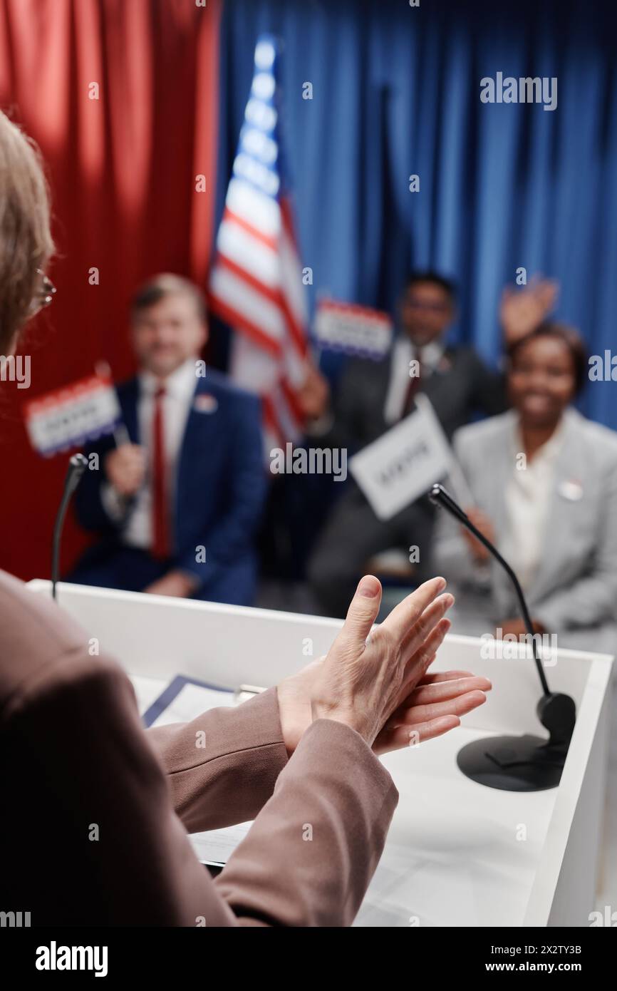 Donna politica che fa un gesto convincente sulla piattaforma mentre parla di fronte a un pubblico interculturale che tiene bandiere di voto alla conferenza Foto Stock
