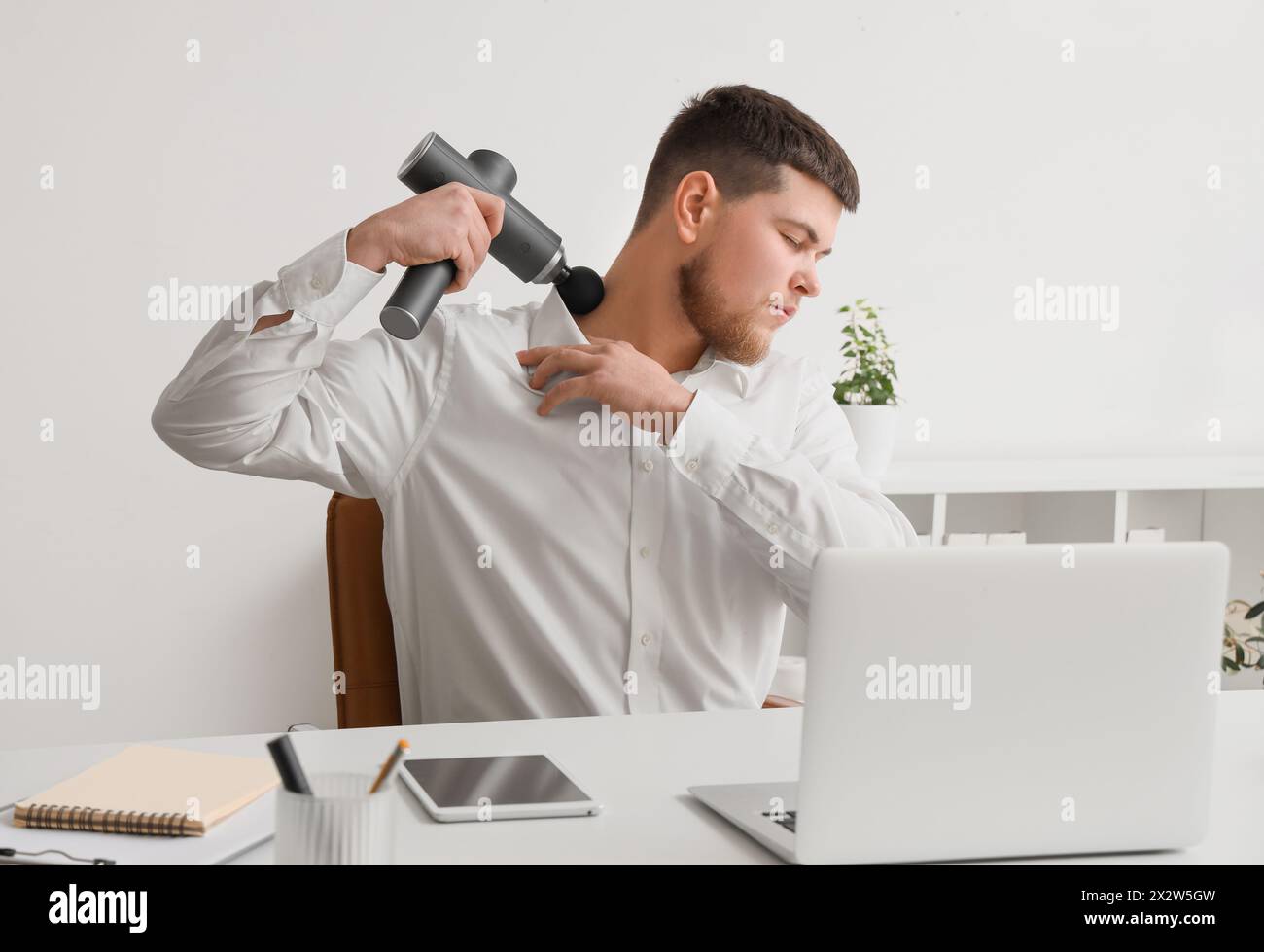 Giovane uomo d'affari che massaggia il collo con un massaggiatore percussivo al tavolo in ufficio Foto Stock