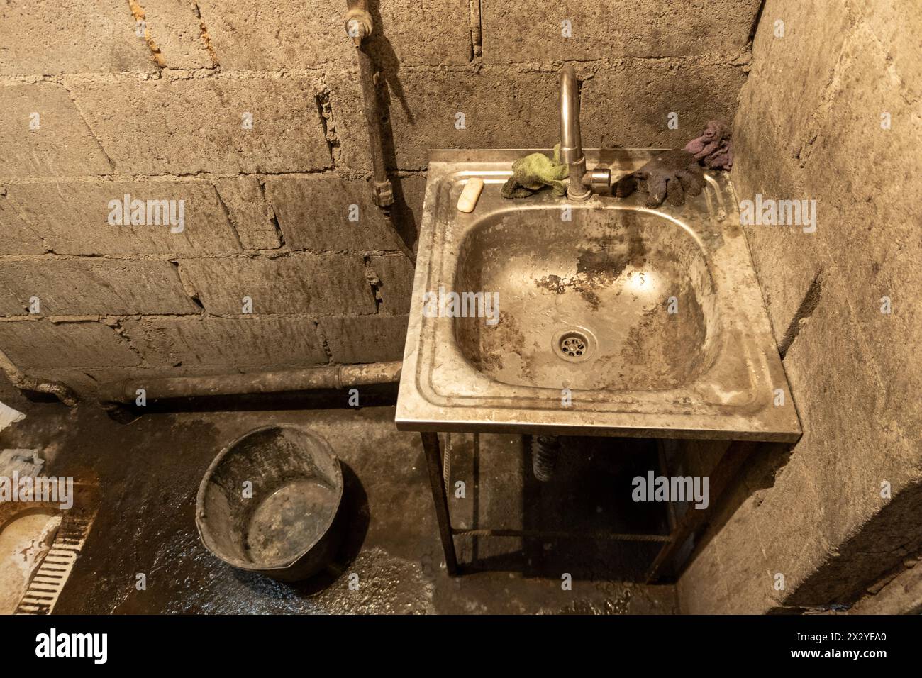 Lavandino in acciaio inossidabile della toilette sporca con secchio sul pavimento, pavimento in cemento non rivestito e pareti in mattoni di cenere. Foto Stock