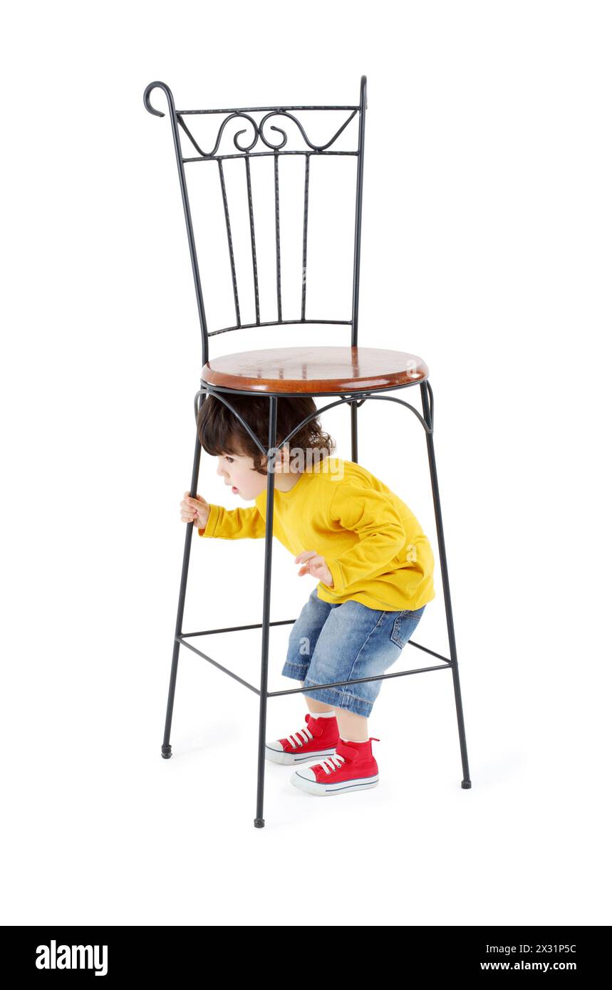 Il bambino si nasconde sotto un'alta sedia in ferro battuto isolata su sfondo bianco. Foto Stock