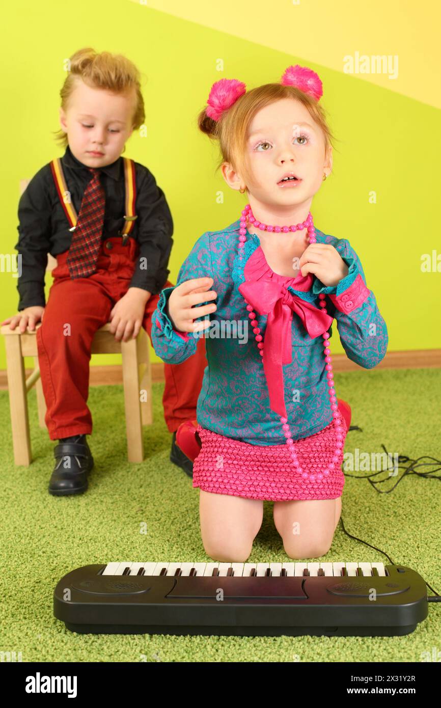 Una bambina carina vicino al pianoforte dei bambini e un ragazzo su una sedia dietro di lei Foto Stock