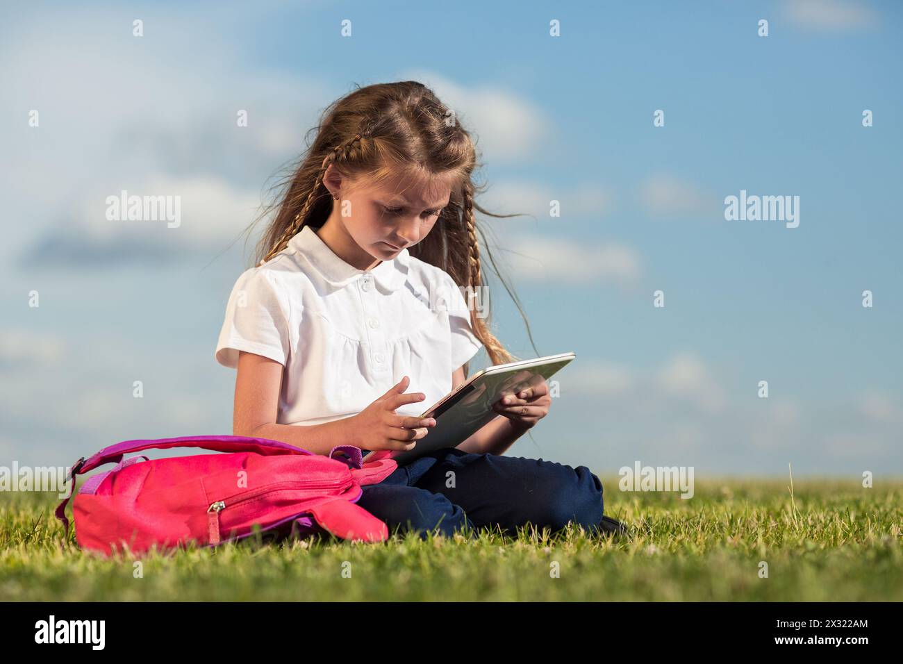 Bambina con lo zaino rosa seduta sull'erba e che guarda in un tablet Foto Stock