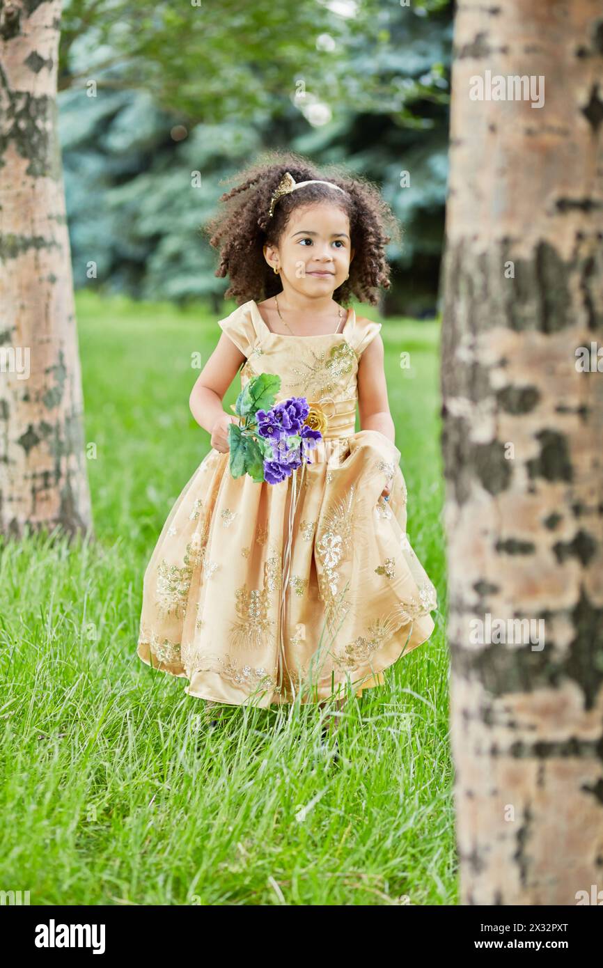 La piccola mulatta in beige party frock si trova tra gli alberi nel parco Foto Stock