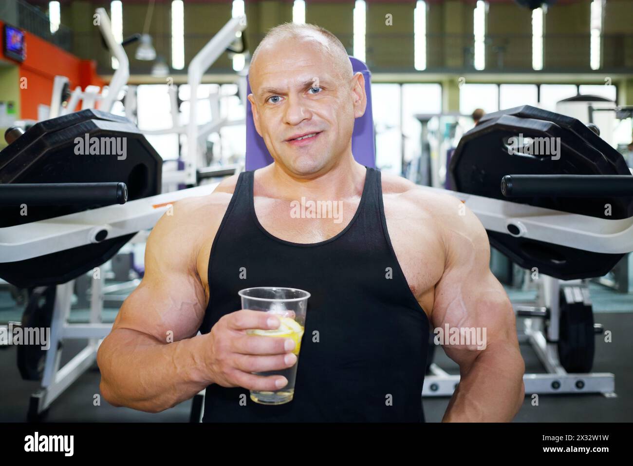 Il bodybuilder in Jersey nero siede sulla macchina per l'esercizio fisico e tiene il bicchiere con acqua al limone nella palestra Foto Stock