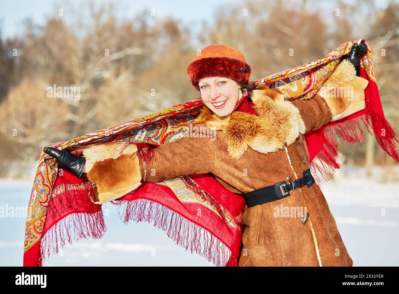 Ritratto di una giovane donna sorridente in pelliccia corta di montone con scialle colorate dietro la schiena contro una foresta invernale Foto Stock