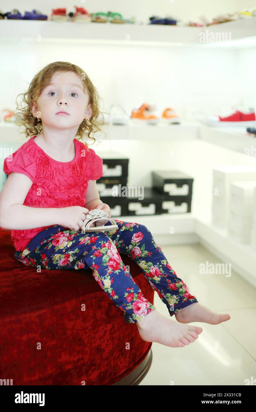 La bambina si siede su un grande pouf e tiene i sandali nel negozio per bambini. Foto Stock