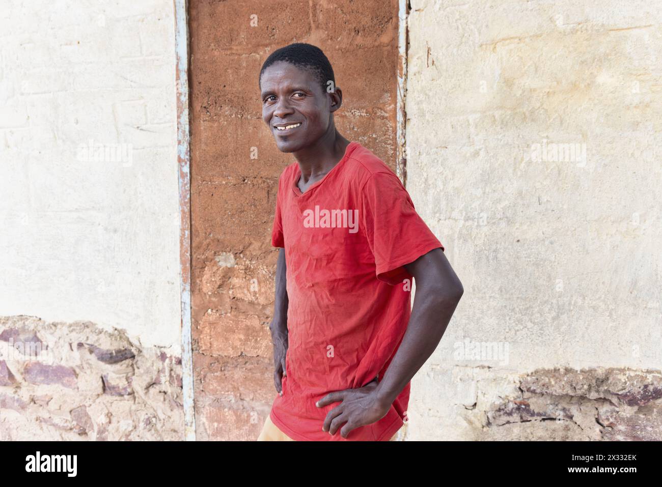 vista del profilo di un giovane ragazzo africano felice, situato di fronte alla casa nel cortile, con indosso una maglietta rossa Foto Stock