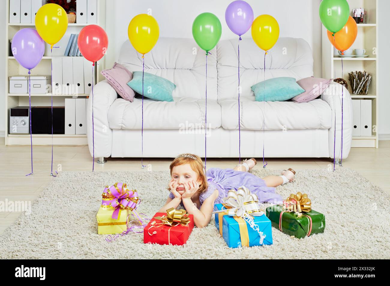 Una bambina vestita con abiti da vacanza giace su un tappeto tra scatole regalo e palloncini di compleanno che si fanno avanti con i suoi hahds Foto Stock