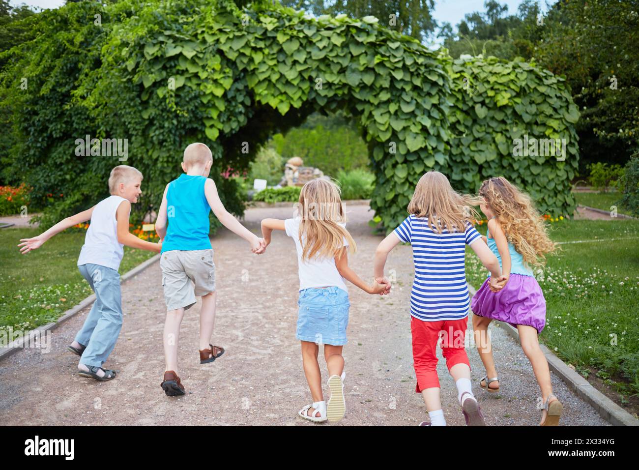 I bambini corrono tenendo le mani sul passaggio pedonale del parco in un arco fatto di piante verdi, vista posteriore, concentrandosi sulle ragazze al centro Foto Stock