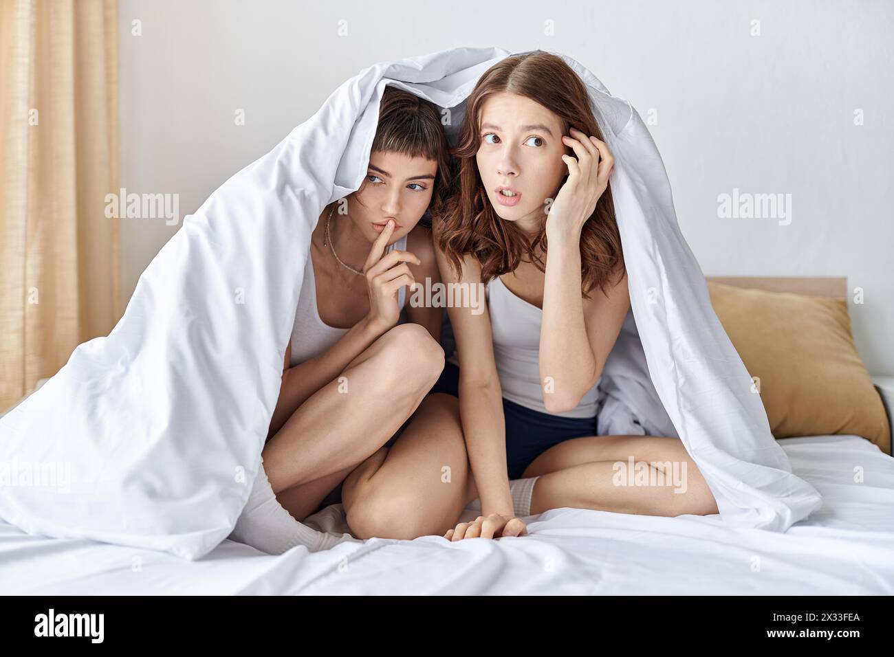 Seduti sotto una coperta su un letto, due donne in abiti comodi condividono un momento tenero. Foto Stock