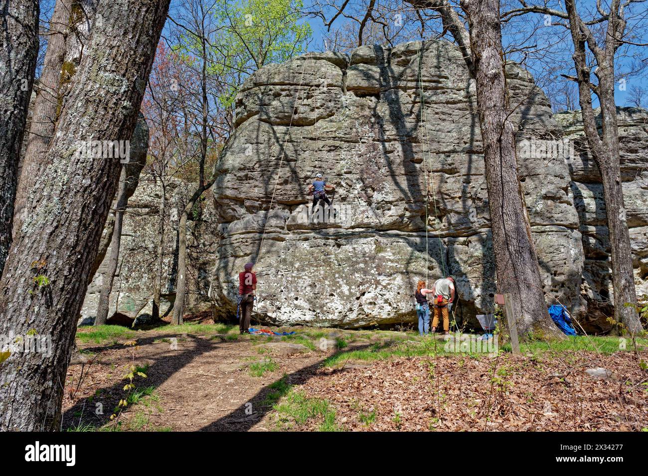 Un arrampicatore che sale sul fianco di un enorme masso con un osservatore che controlla la corda e una coppia che indossa l'attrezzatura per arrampicarsi accanto al sole Foto Stock