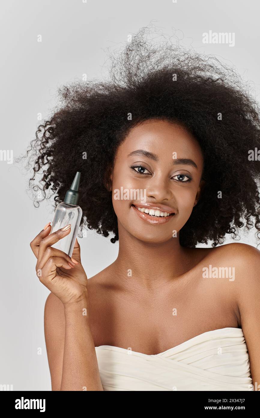 Una splendida giovane donna afroamericana con capelli ricci che presenta una bottiglia di prodotto per capelli in un ambiente vivido da studio. Foto Stock