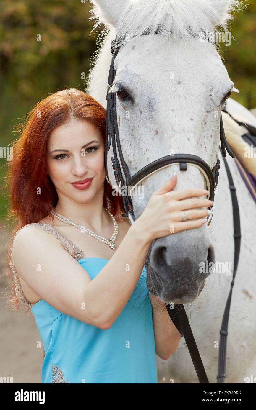 La giovane donna sorridente dai capelli rossi si erge con un cavallo bianco nel parco. Foto Stock