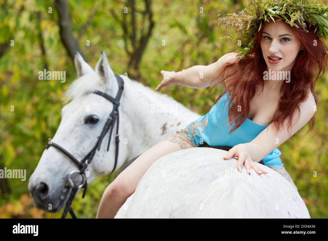 Giovane donna sorridente dai capelli rossi con corona floreale sulla testa siede all'indietro a cavallo nel parco. Foto Stock