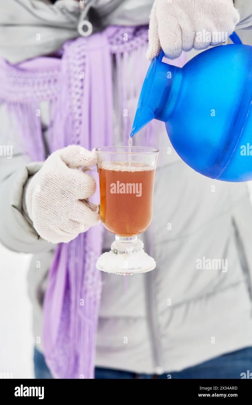 Le mani femminili con guanti bianchi in maglia versano la bevanda calda in una tazza di vetro all'aperto. Foto Stock