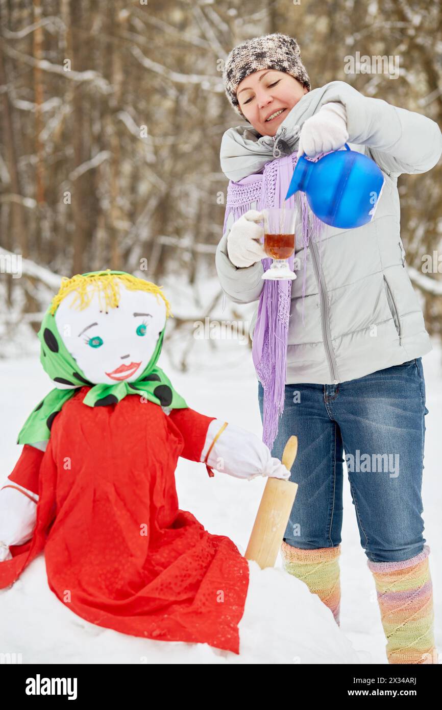 La donna versa la bevanda calda dalla caraffa al vetro in piedi vicino a Maslenitsa ripiena nel parco invernale. Foto Stock