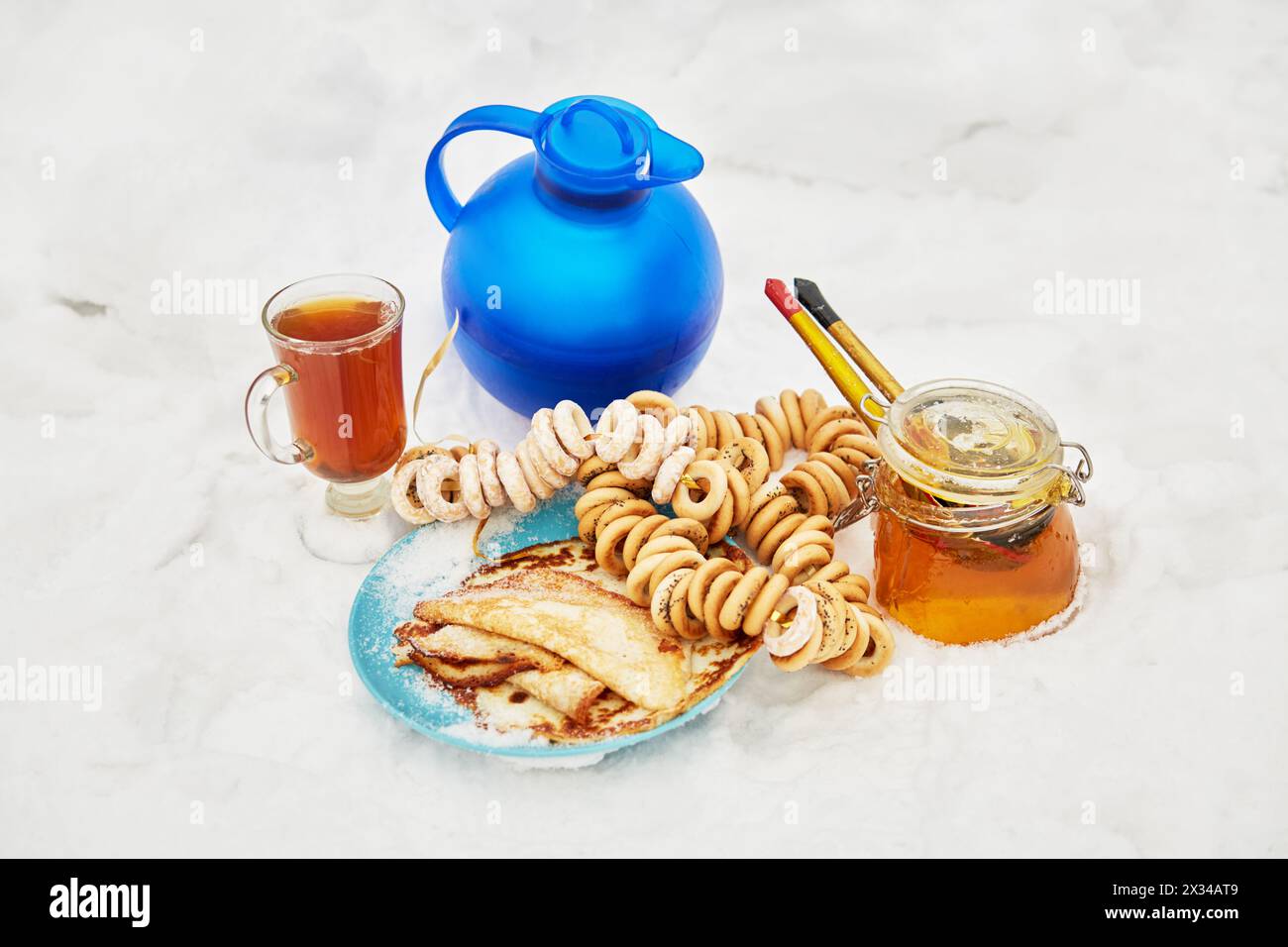 Caraffa blu e tazza in vetro con bevanda, fasci di crackel, vaso di miele, piatto con frittelle sulla neve. Foto Stock
