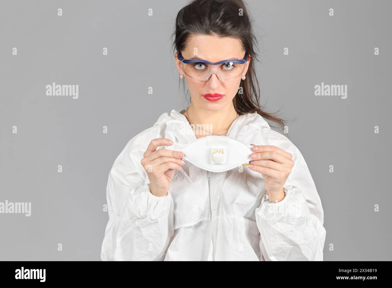 Ritratto di mezza crescita di donna in tuta protettiva bianca di carta sintetica e occhiali, che ci guarda, tiene respiratoria mezza maschera in mano, su ba grigia Foto Stock