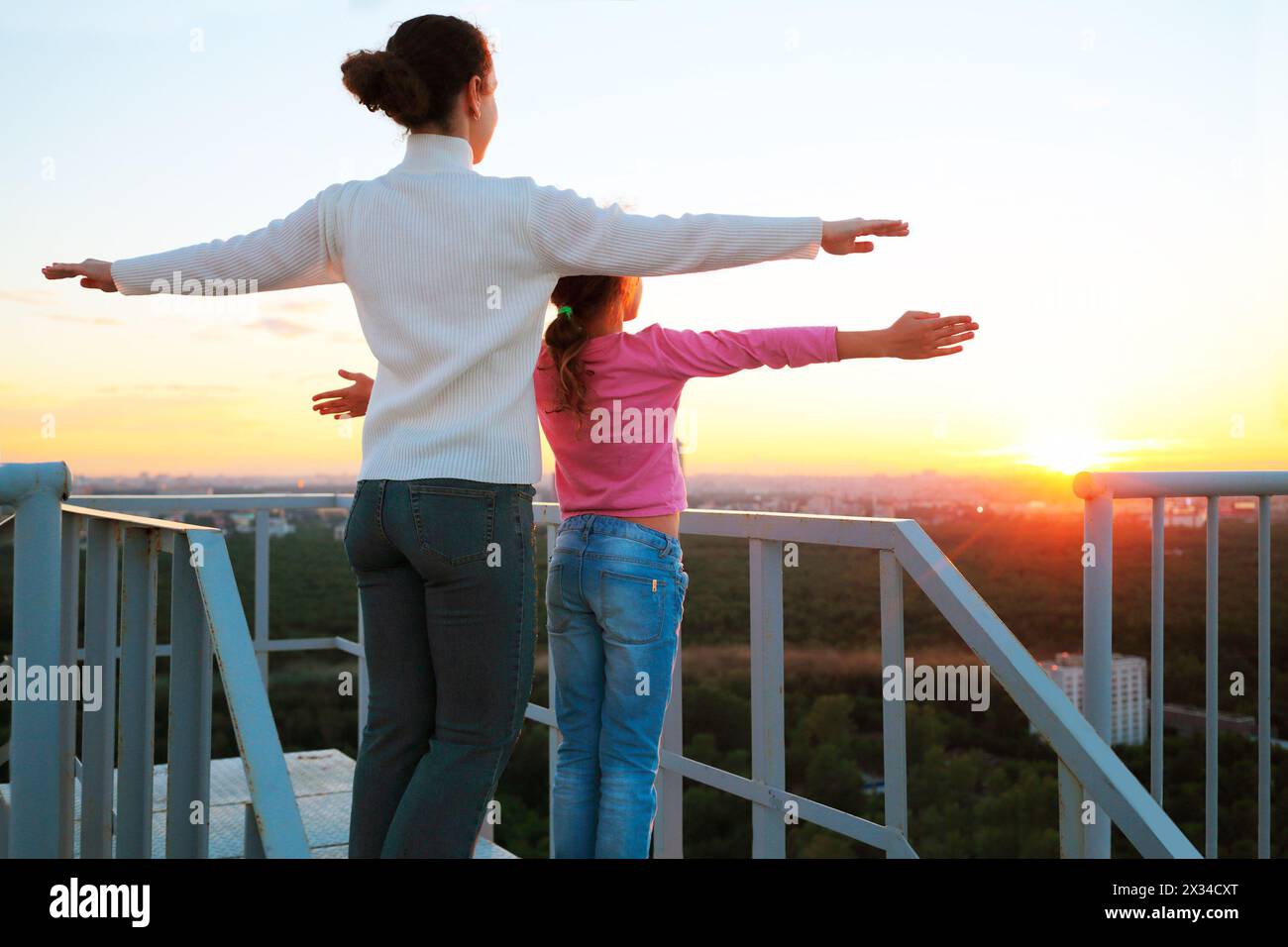 madre e figlia in piedi con le braccia allungate ai lati sul tetto di un edificio a più piani in serata e guardano al tramonto. Foto Stock