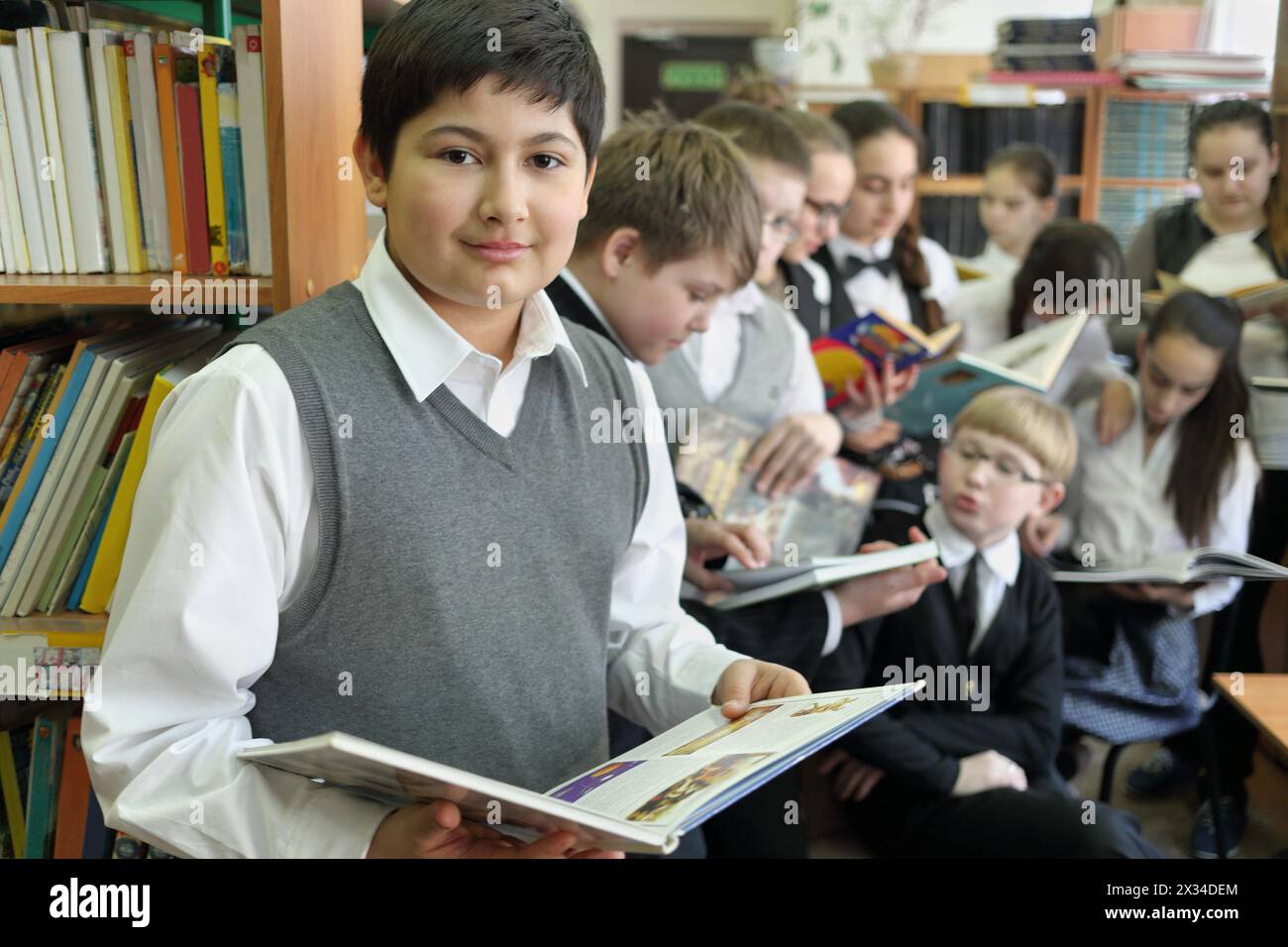 Dieci studenti della biblioteca scolastica che leggono libri sullo sfondo di scaffali con libri, uno in primo piano che guarda in macchina fotografica, si concentrano sul ragazzo a sinistra Foto Stock