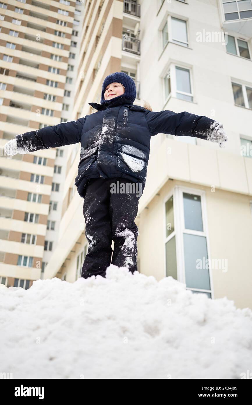 Ragazzo felice, tutto sulla neve, si erge sulla cima del mucchio di neve nel cortile. Foto Stock