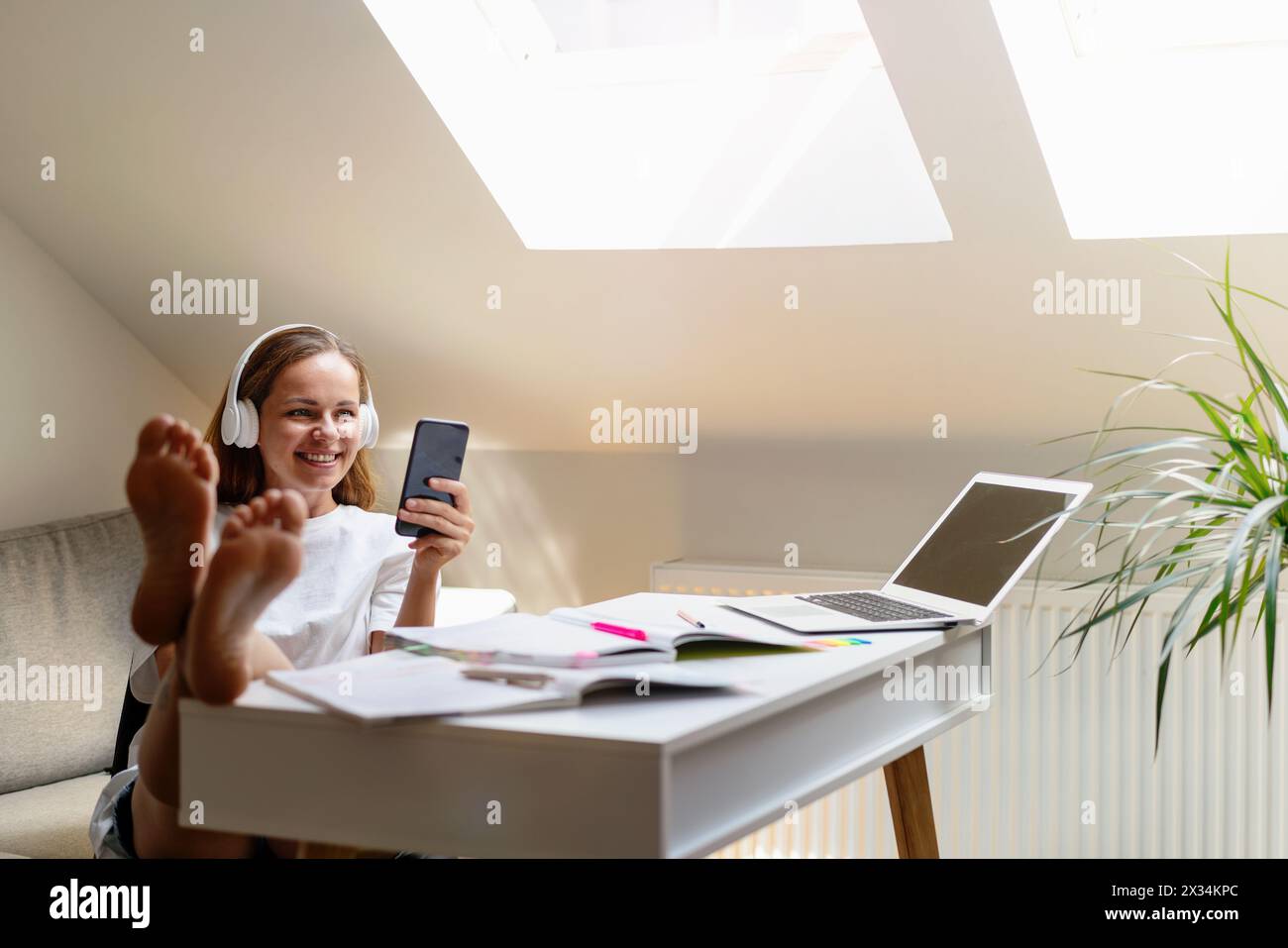 Una studentessa spensierata si siede alla scrivania di casa e chatta online utilizzando il suo smartphone. Foto Stock