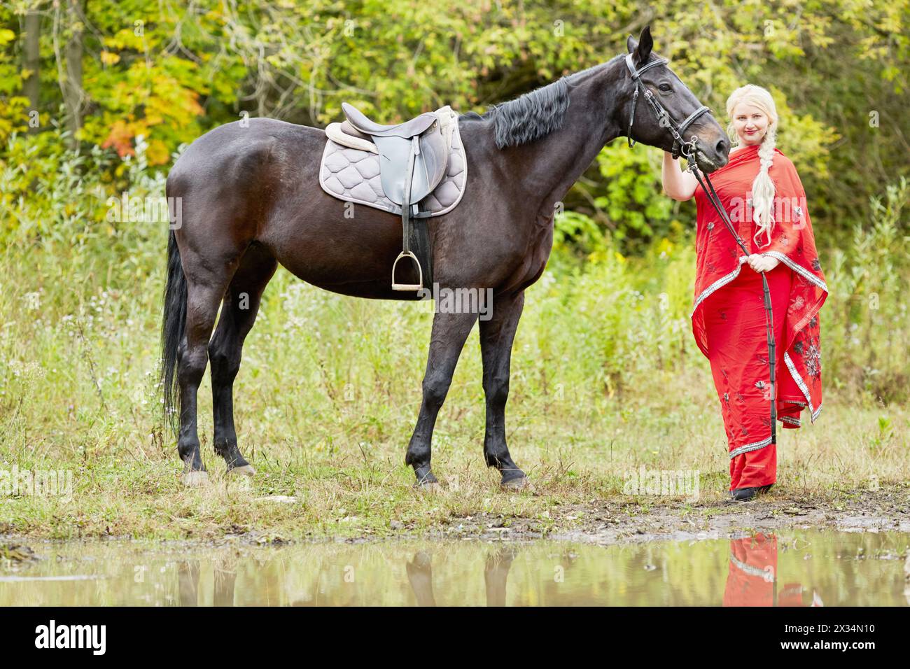 Donna bionda con treccia in abiti rossi si stende con cavallo nel parco vicino all'acqua. Foto Stock
