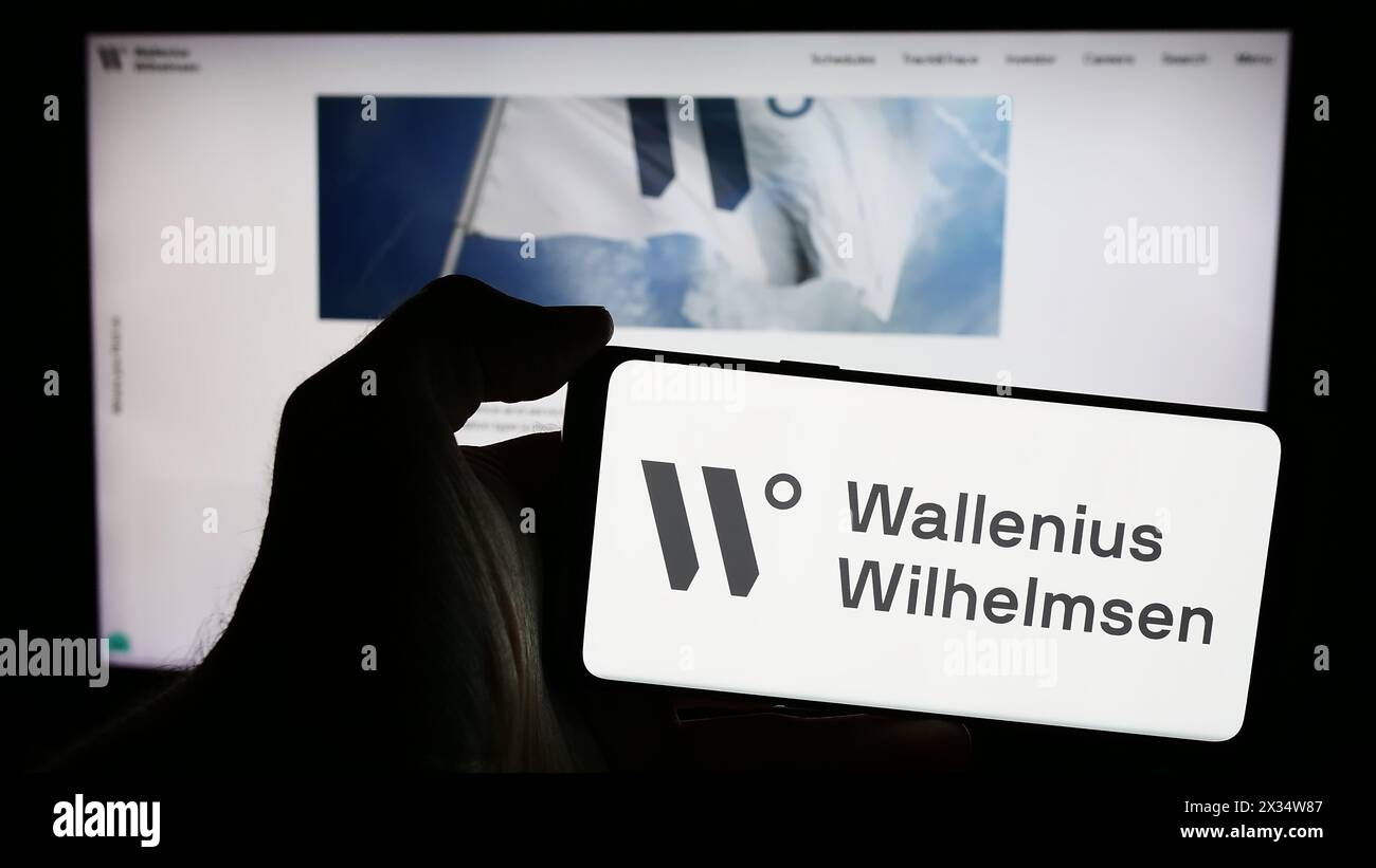 Persona che possiede un telefono cellulare con il logo della compagnia di navigazione Wallenius Wilhelmsen ASA davanti alla pagina web aziendale. Mettere a fuoco il display del telefono. Foto Stock