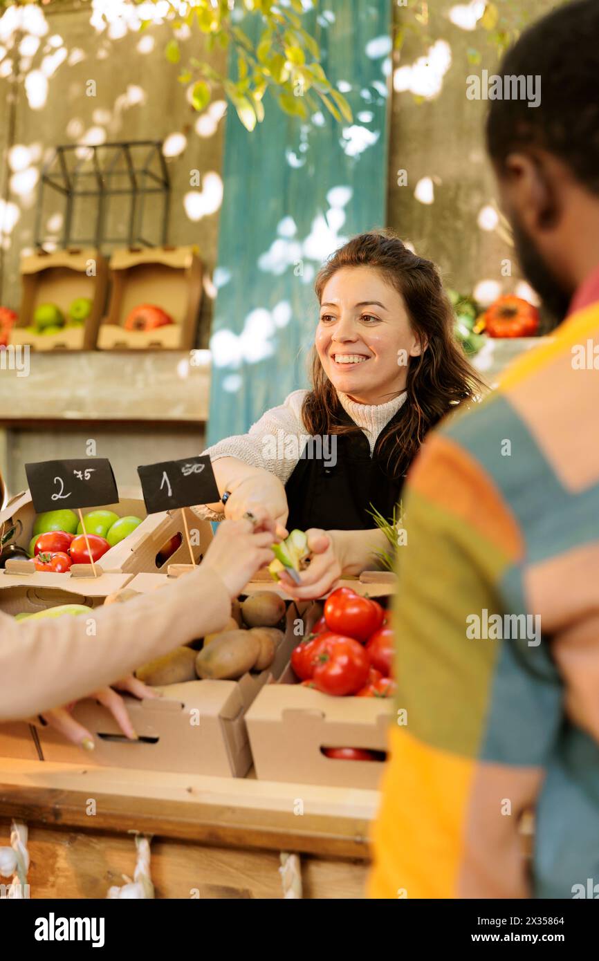 Donna sorridente che offre al cliente la possibilità di provare un piccolo pezzo di mela biologica mentre vende frutta e verdura fresca naturale al festival della fiera del raccolto, attenzione selettiva. Degustazione durante lo shopping. Foto Stock