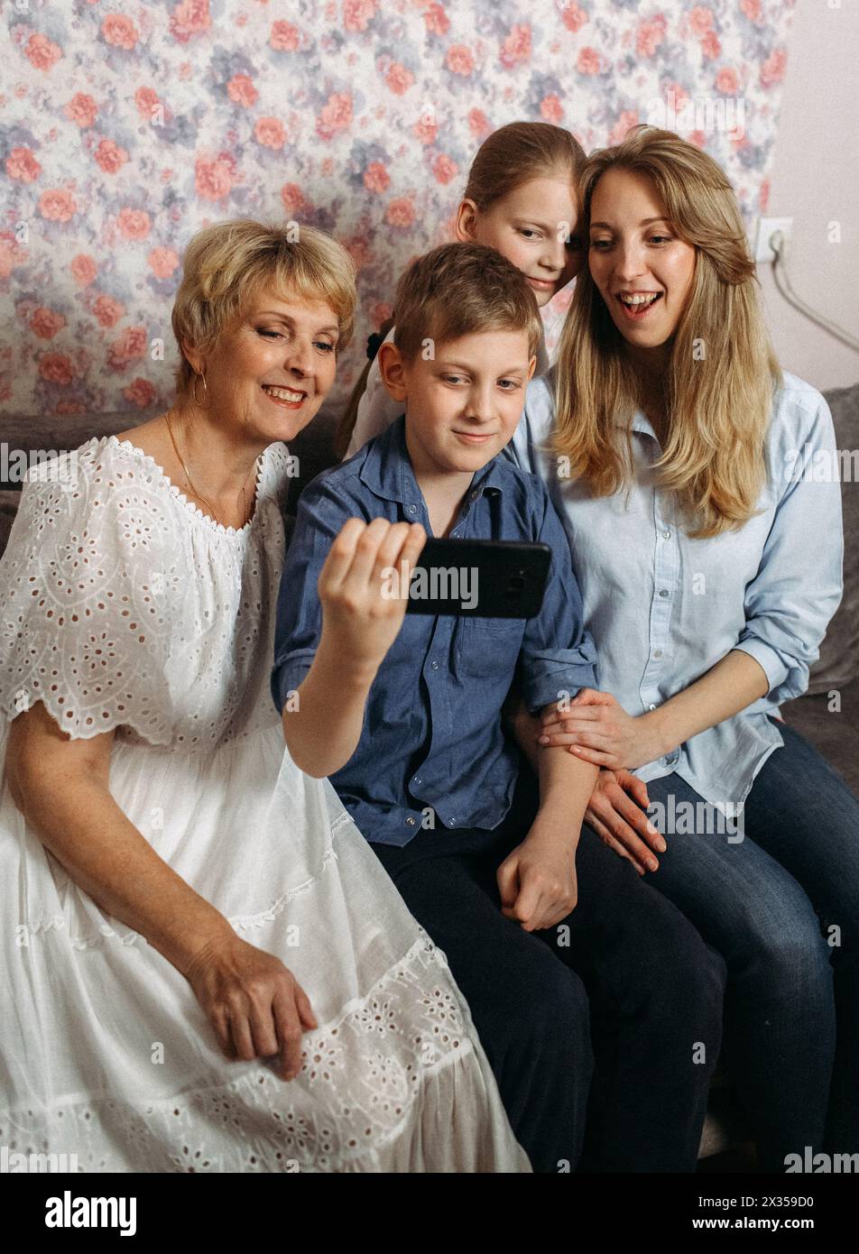 Una famiglia di quattro persone seduta su un divano, sorridente e in posa insieme mentre scatta un selfie con uno smartphone. Foto Stock