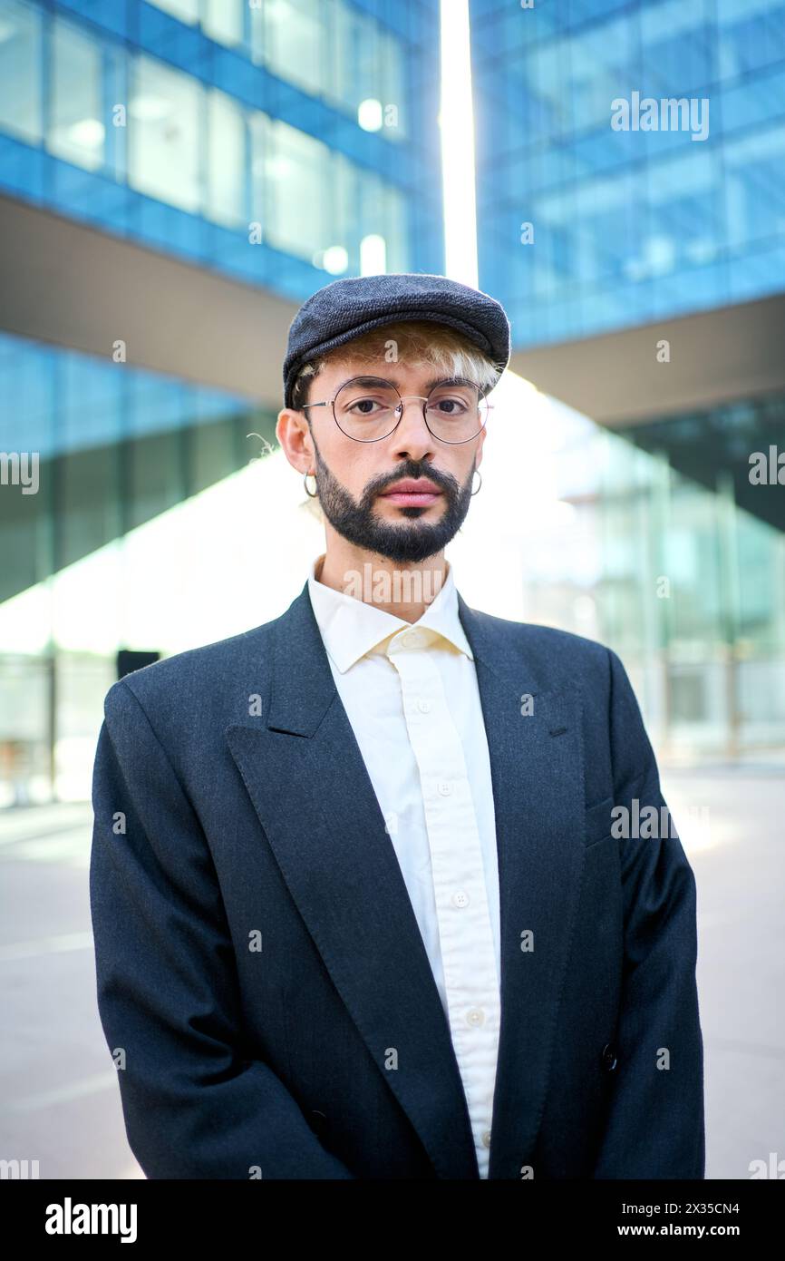 Un giovane hipster caucasico con un'elegante tuta blu, cappello e occhiali in piedi seri e sicuri di sé Foto Stock
