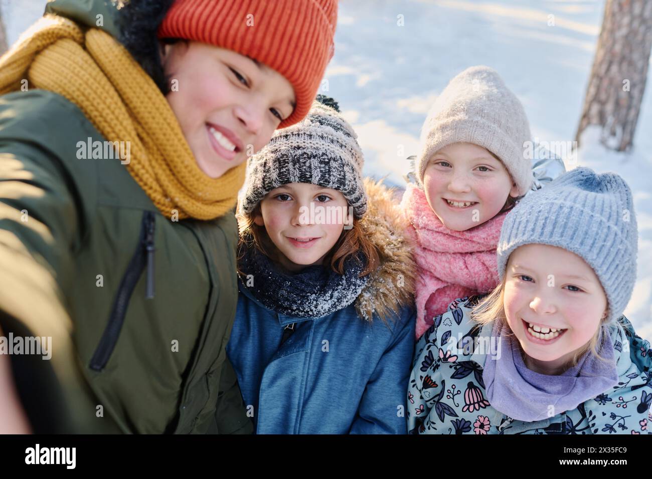 Uno dei quattro bambini felici che scattano selfie sullo smartphone mentre si trova accanto ai suoi allegri amici indossando caldi indumenti invernali Foto Stock