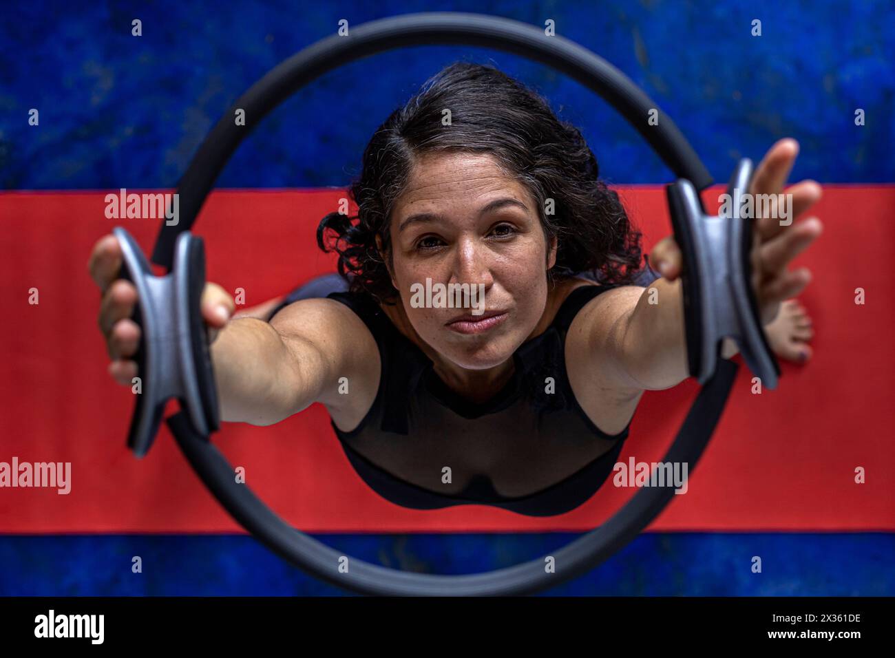 Vista dall'alto della donna latinoamericana (38) che fa esercizi tonificanti con l'anello di esercizio sul tappetino rosso per il fitness. Fondo blu. Studio yoga Foto Stock