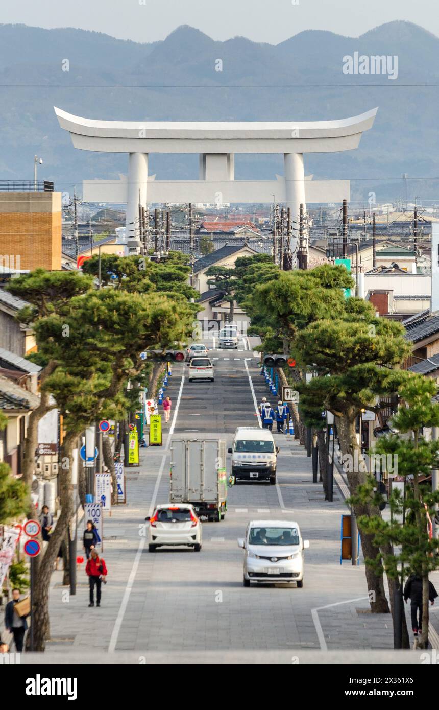 Una strada trafficata con un grande edificio bianco sullo sfondo. La strada è costeggiata da alberi e ha un mix di auto e camion. Foto Stock