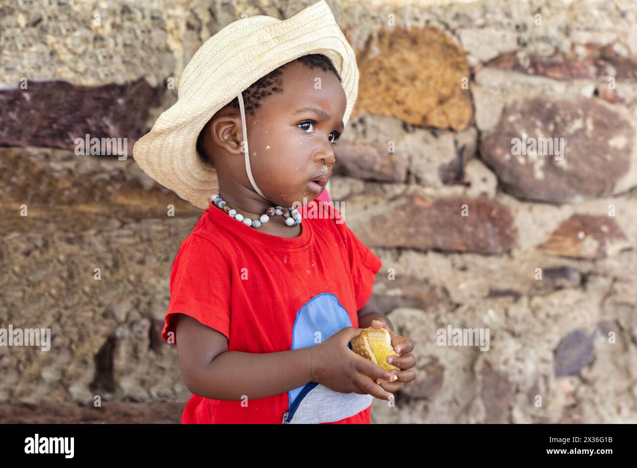 piccola ragazza africana affamata che mangia biscotti davanti alla casa nel cortile, indossando una maglietta rossa e un cappello di paglia Foto Stock
