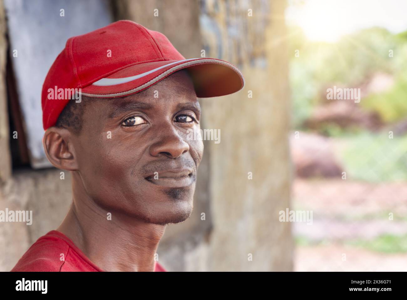 felice uomo africano del villaggio, in piedi all'aperto di fronte alla casa nel cortile al tramonto, indossa un berretto rosso Foto Stock