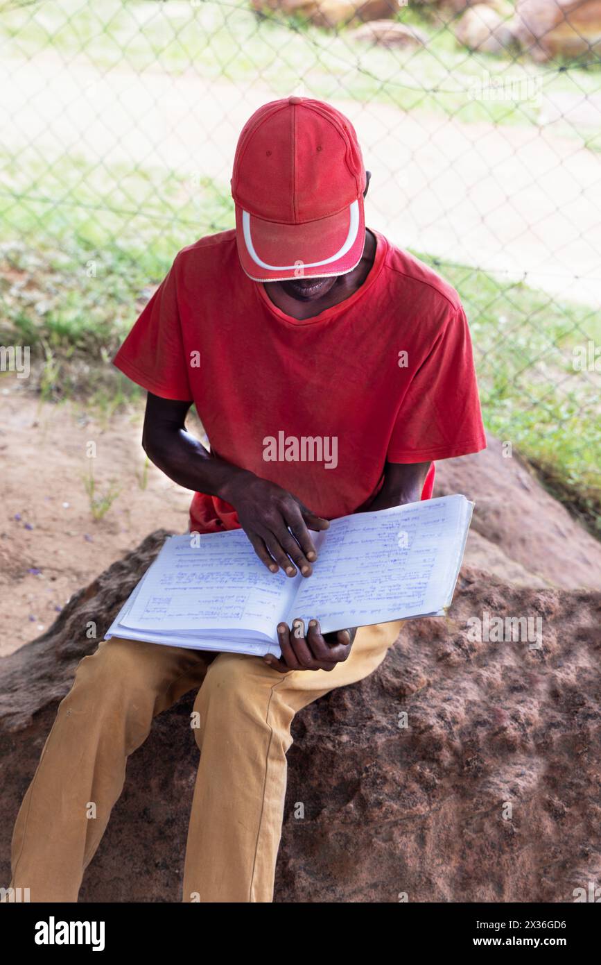 uomo africano del villaggio, che legge la calligrafia da un quaderno di carta, si trova all'aperto su una roccia di fronte alla casa nel cortile Foto Stock