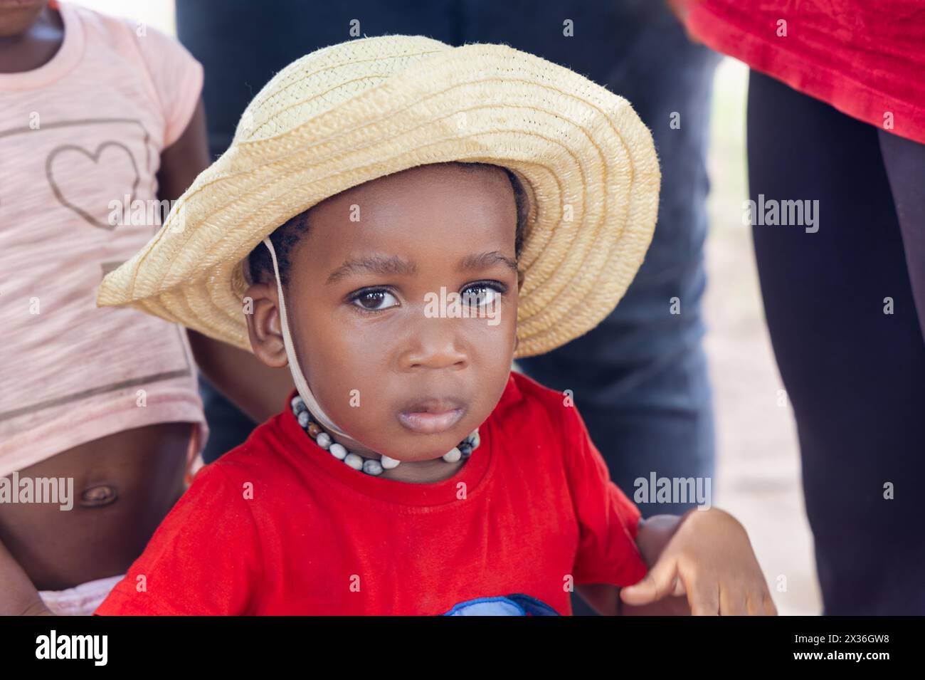 piccola bambina africana di fronte alla casa nel cortile, con una maglietta rossa e un cappello di paglia Foto Stock